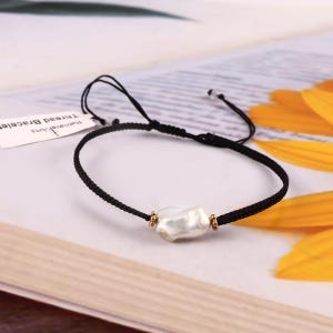 May include: A black thread bracelet featuring a single irregular white pearl at the center, flanked by two small gold-toned beads. The bracelet has adjustable knotted ends and a white paper tag with the text Ratnavali Arts Thread Bracelet.