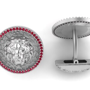 Può includere: Gemelli in argento con un design dettagliato a testa di leone. La testa del leone è circondata da un bordo strutturato e da un anello di piccole pietre preziose rosse. Chiusura classica.