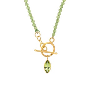 May include: A gold-toned necklace with a toggle clasp and a green gemstone pendant. The necklace features a strand of small, faceted green beads. The pendant is a marquise-shaped, light green gemstone set in a gold frame.