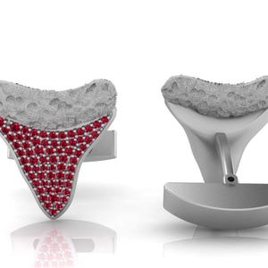 Può includere: Un paio di gemelli a forma di denti di squalo. Un gemello presenta una sezione superiore grigia testurizzata e una parte triangolare inferiore con pavé di numerose piccole pietre rosse. L'altro mostra il retro in metallo grigio liscio e solido.