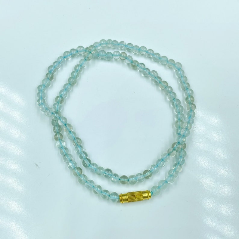 May include: A light blue gemstone necklace with a gold clasp. The necklace is made of round beads and is approximately 18 inches long.