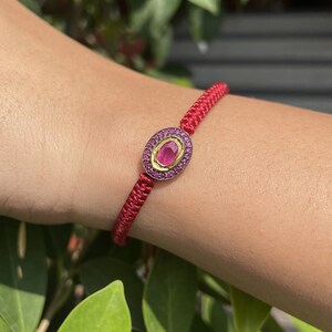 May include: A red cord bracelet featuring a central oval-shaped charm. The charm has a gold-toned inner border surrounding a single pink gemstone, encircled by a dark metal frame embedded with numerous small, round, vibrant pink stones.