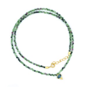 May include: A beaded necklace featuring small, faceted green and purple stones strung together. The necklace includes a gold-toned metal chain extender with a lobster clasp and a single oval blue stone pendant hanging from the bottom of the strand.