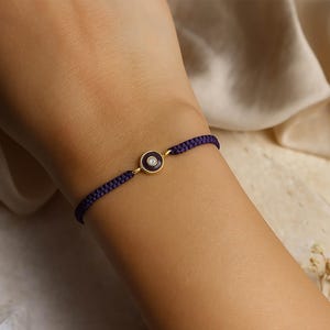 Könnte beinhalten: Ein Armband aus dunkelviolettem geflochtenem Kordelmaterial mit einem zentralen, kreisförmigen, goldfarbenen Anhänger. Der Anhänger besitzt eine dunkelviolette Emaille-Einlage mit einem kleinen, klaren, runden Edelstein in der Mitte.