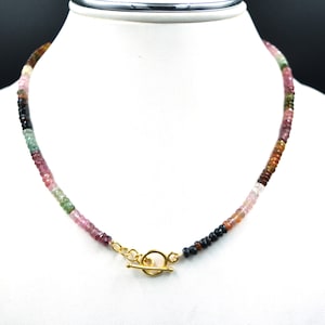 Op de afbeelding: Een ketting bestaande uit kleine, gefacetteerde, veelkleurige toermalijnkralen in tinten roze, groen, bruin en zwart. De streng is voorzien van een goudkleurige metalen knevelsluiting bestaande uit een ronde ring en een T-balk in het midden.