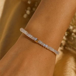 Op de afbeelding: Een delicate armband bestaande uit kleine, doorschijnende, gefacetteerde kralen met een subtiele iriserende glans. De streng bevat een enkele, gladde, cilindrische goudkleurige kraal nabij het midden, wat een verfijnd en eenvoudig accessoire vormt.