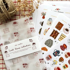 Puede incluir: Un conjunto de hojas de pegatinas titulado Sweet Home Collection con ilustraciones de tazas, libros, un suéter, un cartón de leche y arreglos florales. Las pegatinas vienen en un sobre translúcido con una banda de papel con el texto impreso.