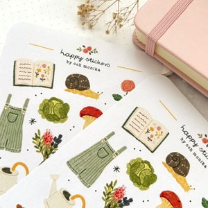 Pu&ograve; includere: Un foglio di adesivi a tema giardino. Gli adesivi presentano illustrazioni di un libro, una salopette, una lumaca, un fungo, un cavolo, un annaffiatoio e fiori. Il testo "happy stickers by ooch monika" &egrave; stampato sul foglio.