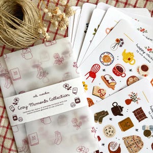 May include: A collection of stickers and decorative paper featuring cozy-themed illustrations. The stickers showcase images of tea, books, and baked goods. The paper is wrapped around the stickers and has a repeating pattern of cozy objects. The text "Cozy Moments Collection" is visible.