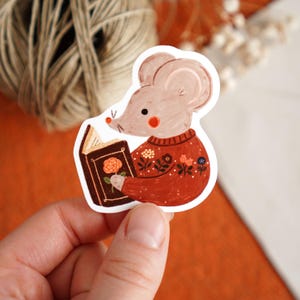 May include: A sticker featuring a cartoon mouse reading a book. The mouse wears a rust-colored sweater with floral details. The book has a brown cover with a rose illustration. The mouse has pink cheeks and a small nose.