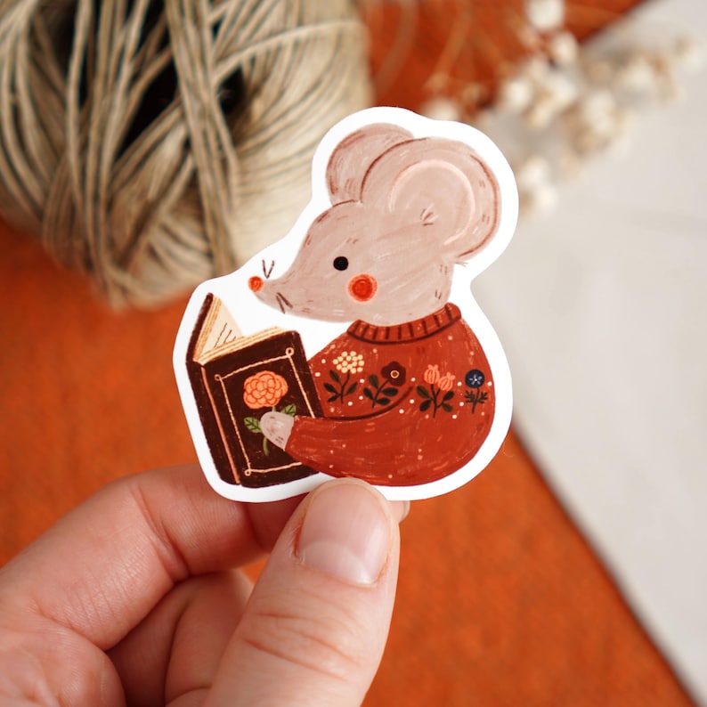 May include: A sticker featuring a cartoon mouse reading a book. The mouse wears a rust-colored sweater with floral details. The book has a brown cover with a rose illustration. The mouse has pink cheeks and a small nose.