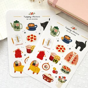 Op de afbeelding: Twee vellen met stickers met verschillende illustraties, waaronder een theepot, een kopje thee, een kat, een gebreide sjaal, een pot jam, een stoel, een kom met koekjes, een ovenschaal, een bloempot en een kruiswoordpuzzel. De stickers zijn gelabeld met "happy stickers by ooch monika".
