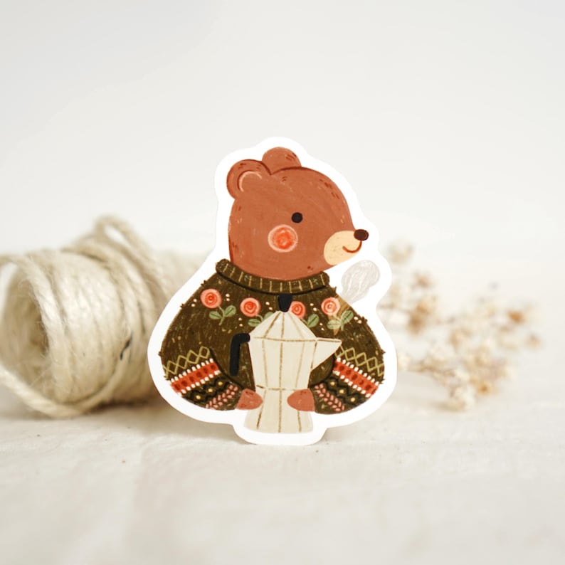 K&ouml;nnte beinhalten: Ein Aufkleber mit einem braunen B&auml;ren, der einen gr&uuml;nen Pullover mit floralen und geometrischen Mustern tr&auml;gt. Der B&auml;r h&auml;lt einen wei&szlig;en Kaffeezubereiter. Dampf steigt aus dem Topf auf, was auf frisch gebr&uuml;hten Kaffee hindeutet. Der Aufkleber hat einen skurrilen, illustrierten Stil.