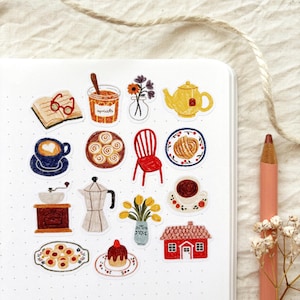 May include: A collection of illustrated stickers on a white notebook. The stickers depict various cozy items, including a cup of coffee, a teapot, pastries, a red chair, and a small house. A pencil and dried flowers are in the background.