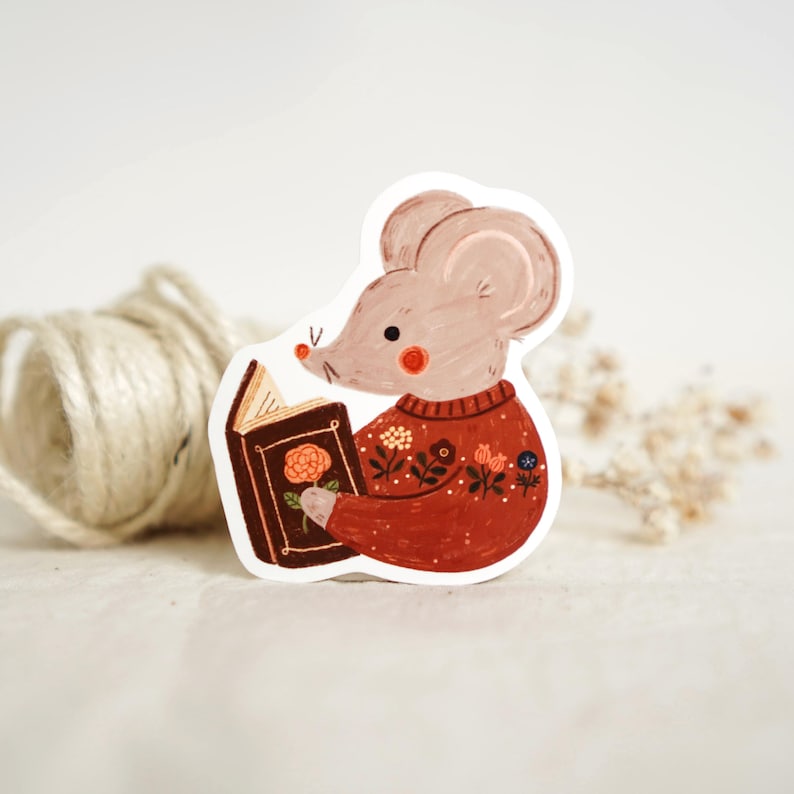 May include: A sticker featuring a cartoon mouse reading a book. The mouse is wearing a red sweater with floral details. The book is brown with a rose illustration on the cover. The mouse has pink cheeks and a small black nose.