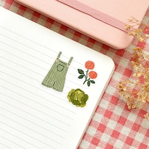 Pu&ograve; includere: Tre adesivi illustrati su un taccuino bianco a righe. Gli adesivi sono di una salopette verde, un cavolo verde e una rosa rossa con uno stelo e foglie verdi.