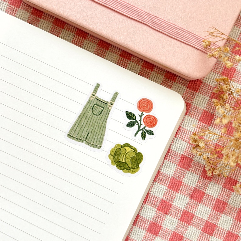 Pu&ograve; includere: Tre adesivi illustrati su un taccuino bianco a righe. Gli adesivi sono di una salopette verde, un cavolo verde e una rosa rossa con uno stelo e foglie verdi.