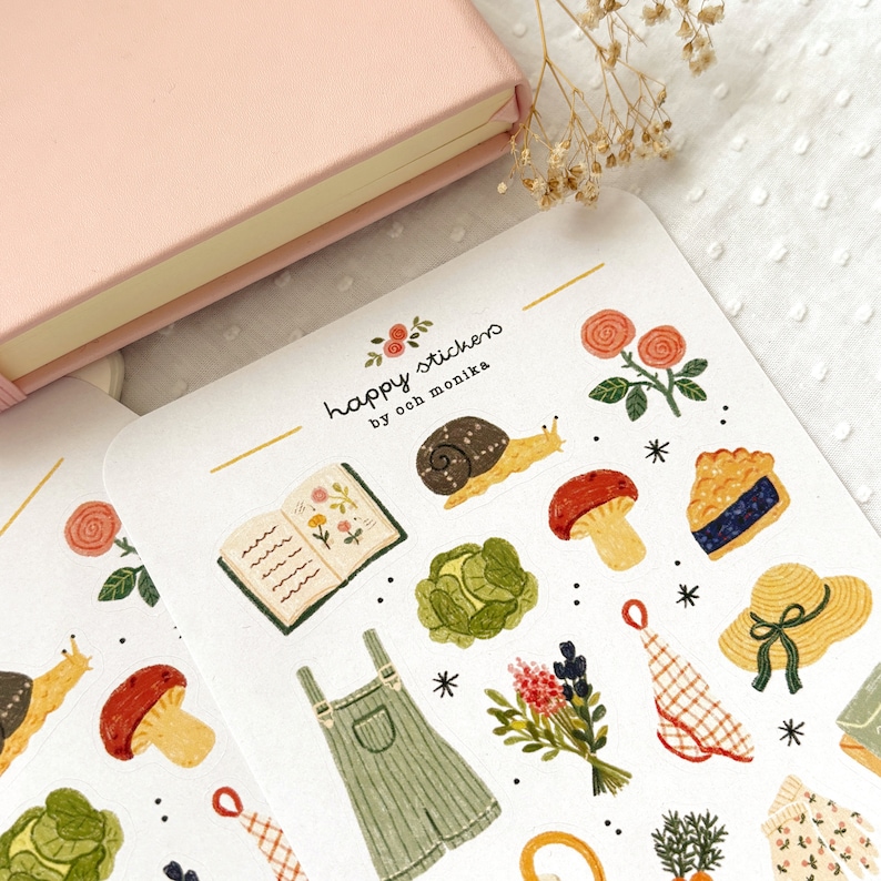 Pu&ograve; includere: Un foglio di adesivi con una variet&agrave; di illustrazioni, tra cui una lumaca, un fungo, un libro, un cavolo, un mazzo di fiori, una salopette, una torta, un cappello, uno strofinaccio e un rametto di rosmarino. Gli adesivi sono su uno sfondo bianco e hanno un bordo dorato. Il testo "happy stickers by ooch monika" &egrave; stampato nella parte superiore del foglio.