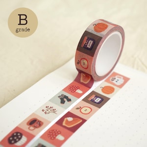 May include: A roll of decorative washi tape featuring a repeating pattern of autumn-themed illustrations including pumpkins, jam jars, apples, mushrooms, socks, and mugs. The tape is partially unrolled, displaying the colorful square icons on a white background.