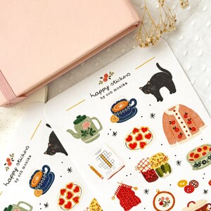 Op de afbeelding: Een vel stickers met illustraties van een theepot, een kopje thee, een kat, een trui, een bord met koekjes, een pot jam, een gebreide sjaal, een borduurring, een knoop en een kruiswoordpuzzel. De stickers zijn gelabeld "happy stickers by och monika".