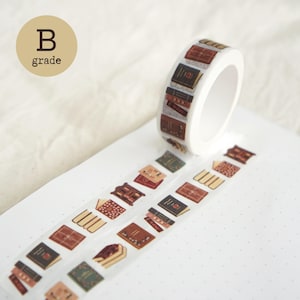 Puede incluir: Un rollo de cinta adhesiva transparente con un patrón repetitivo de varios libros, estanterías y pilas de libros de estilo antiguo en tonos marrones, rojos y verdes. Una etiqueta circular beige en la esquina superior izquierda muestra el texto B grade.