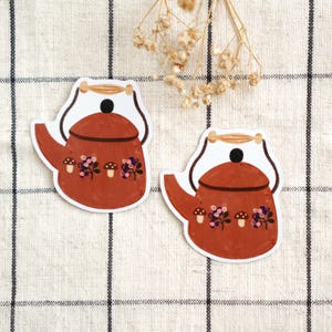 Puede incluir: Dos pegatinas troqueladas que muestran una tetera de color terracota con un asa marrón oscuro, un pomo de tapa negro y un patrón decorativo de pequeñas setas marrones y racimos de bayas azules en la parte frontal de la tetera.
