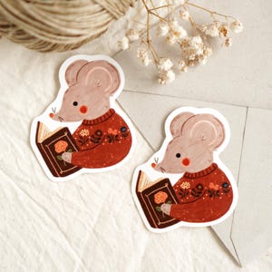 May include: Two stickers featuring a cartoon mouse wearing a red sweater with floral details, reading a book with a rose on the cover. The mouse has a light brown body, pink cheeks, and a small red nose. The stickers are on a white background.