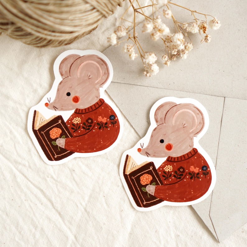May include: Two stickers featuring a cartoon mouse wearing a red sweater with floral details, reading a book with a rose on the cover. The mouse has a light brown body, pink cheeks, and a small red nose. The stickers are on a white background.