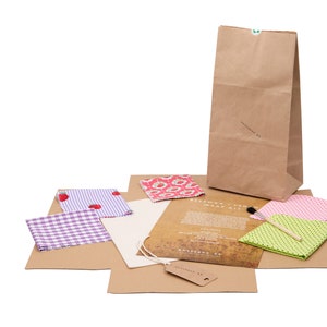May include: A brown paper bag with a green sticker, a set of four fabric squares with different patterns, a paintbrush, and a sheet of instructions. The fabric squares are pink with white polka dots, green with white polka dots, purple with white squares, and pink with red flowers. The instructions are for a beeswax food wrap kit.