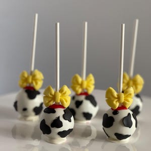 May include: Five cake pops decorated with black and white cow print and yellow bows. The cake pops are on white sticks and are arranged on a white cake stand.