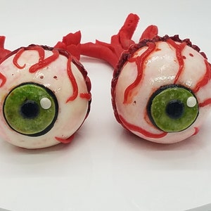 K&ouml;nnte beinhalten: Zwei realistisch aussehende Augen-Cake-Pops mit rotem Zuckerguss, der Blutgef&auml;&szlig;e simuliert. Die Augen sind wei&szlig; mit gr&uuml;nen Iris und schwarzen Pupillen.
