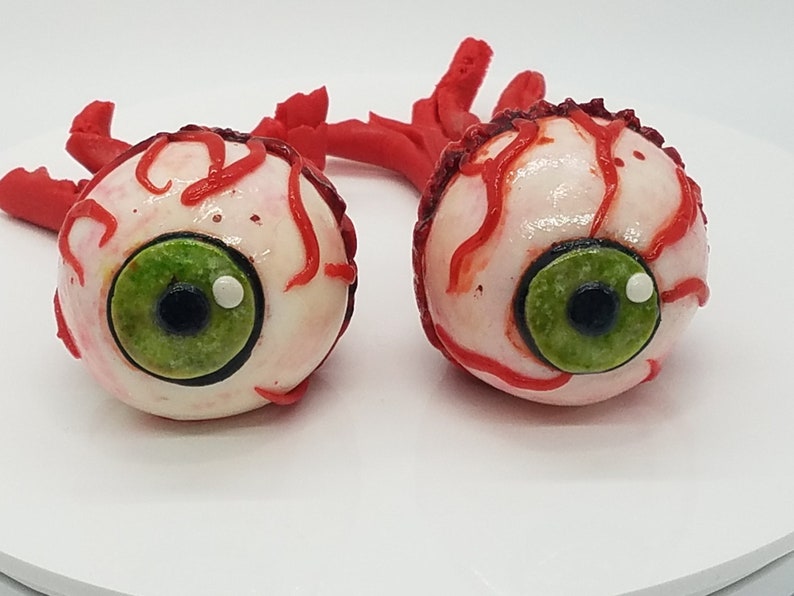 K&ouml;nnte beinhalten: Zwei realistisch aussehende Augen-Cake-Pops mit rotem Zuckerguss, der Blutgef&auml;&szlig;e simuliert. Die Augen sind wei&szlig; mit gr&uuml;nen Iris und schwarzen Pupillen.