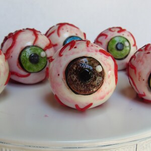 K&ouml;nnte beinhalten: Sechs realistisch aussehende Augen-Cake-Pops mit roten Adern und verschiedenen farbigen Iris. Die Cake-Pops befinden sich auf einem wei&szlig;en Kuchenst&auml;nder.