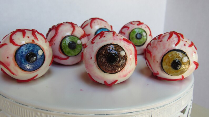 K&ouml;nnte beinhalten: Sechs realistisch aussehende Augen-Cake-Pops mit roten Adern und verschiedenen farbigen Iris. Die Cake-Pops befinden sich auf einem wei&szlig;en Kuchenst&auml;nder.