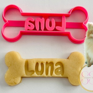 May include: A pink plastic dog bone-shaped cookie cutter with the name "Luna" cut out of a baked cookie. A fluffy, light-colored Pomeranian dog is in the background.