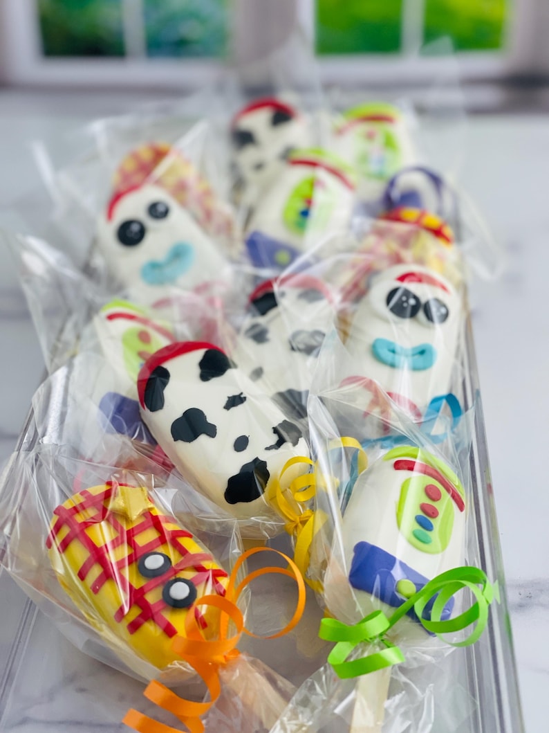 May include: A clear plastic container filled with colorful cake pops decorated with characters from the Toy Story movie franchise. The cake pops are on sticks and wrapped in clear plastic bags with colorful ribbons.