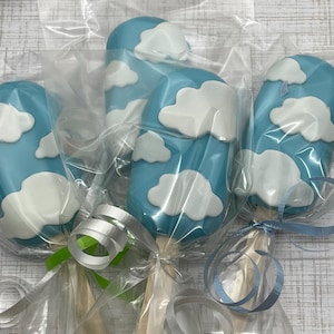May include: Four blue and white cake pops decorated with clouds. The cake pops are individually wrapped in clear plastic and tied with blue and green ribbon.