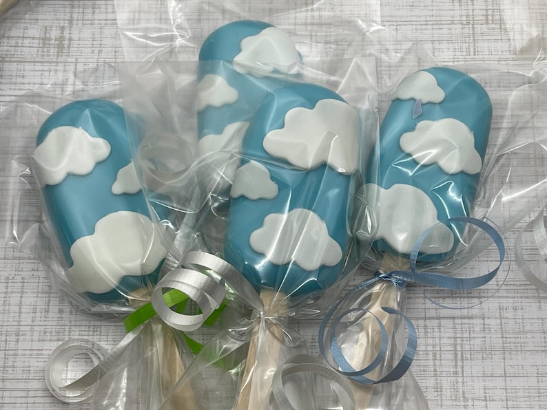 May include: Four blue and white cake pops decorated with clouds. The cake pops are individually wrapped in clear plastic and tied with blue and green ribbon.