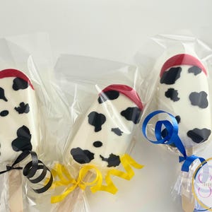 May include: Three white chocolate covered cake pops decorated with a black and white cow print design. Each cake pop has a red top and is wrapped in clear plastic. The cake pops are tied with blue, yellow, and black ribbon.