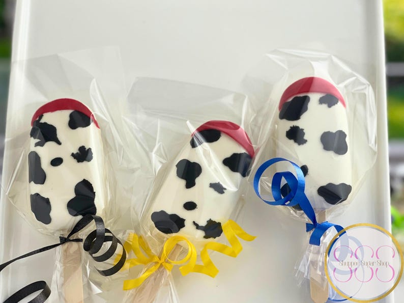 May include: Three white chocolate covered cake pops decorated with a black and white cow print design. Each cake pop has a red top and is wrapped in clear plastic. The cake pops are tied with blue, yellow, and black ribbon.