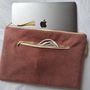 Pode incluir: Uma capa para laptop de veludo cotelê rosa empoeirado com zíper dourado e uma pequena borla. A capa tem um bolso com um cabo de carregamento branco dentro. Um laptop prateado com o logotipo da Apple é parcialmente visível.