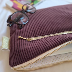 Può includere: Una custodia per laptop in velluto a coste bordeaux con cerniera dorata e tiretto dorato. La fodera interna è bianca con pois dorati. Occhiali tartarugati poggiano sopra la custodia. Un nastro dorato è attaccato al lato.