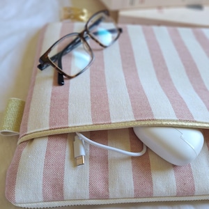 May include: A pink and white striped laptop sleeve with a gold zipper and trim. A pair of eyeglasses rests on top. A white computer mouse and charging cable are visible in the pocket. The text "LNAtelier" is at the bottom.