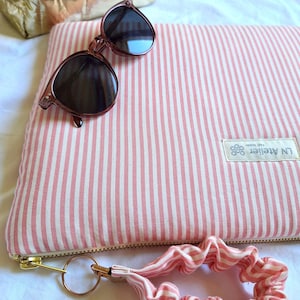 Può includere: Una pochette a righe rosa e bianche con cerniera dorata, uno scrunchie abbinato e occhiali da sole rosa. Sullo sfondo una borsa metallica dorata. La pochette ha un'etichetta con la scritta "LN Atelier".
