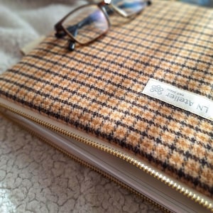 May include: A rectangular laptop sleeve featuring a tan and brown houndstooth pattern with a gold-toned metal zipper. A white fabric label is stitched to the front with the text LN Atelier and Fait Main. A pair of brown-rimmed eyeglasses rests on top.