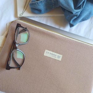 May include: A rectangular laptop sleeve featuring a brown and white micro-check pattern with a gold-toned zip closure. A white fabric label is stitched to the front with the text LN Atelier. A pair of dark brown tortoiseshell glasses rests on top.