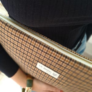 Può includere: Una custodia per laptop a quadri marrone e beige con cerniera dorata. La custodia ha un'etichetta bianca con la scritta "LN Atelier". La custodia è tenuta da una persona che indossa una camicia nera e jeans blu.