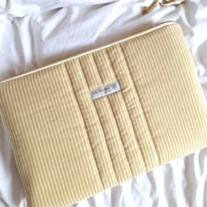 Può includere: Una custodia per laptop a righe gialle e bianche con un design trapuntato. La custodia ha una chiusura a zip e una piccola etichetta con la scritta "LN Atelier" con un logo a forma di farfalla.