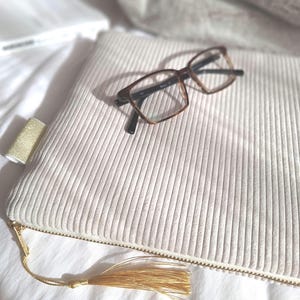 Puede incluir: Un bolso de mano de pana color crema con cremallera y borla doradas, junto con gafas. Las gafas tienen una montura marrón carey. El bolso de mano es rectangular y tiene una textura acanalada vertical.