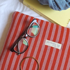 Può includere: Una pochette a righe rosse e bianche con un paio di occhiali e un braccialetto dorato. La pochette ha un'etichetta bianca con la scritta "LN Atelier". Un libro giallo e una giacca di jeans sono sullo sfondo.
